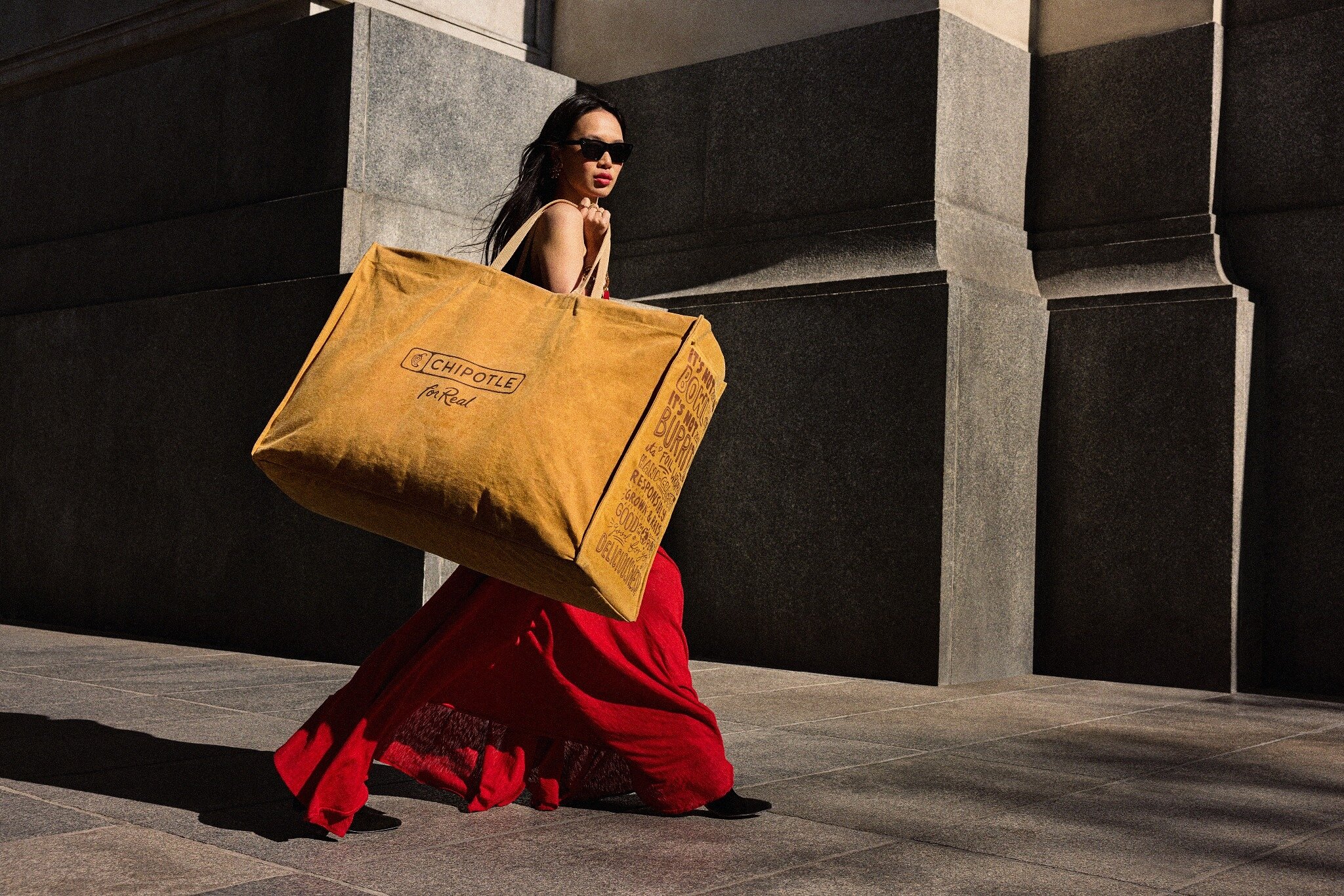 A woman in sunglasses and a red skirt carries an oversized golden Chipotle bag along a city street.