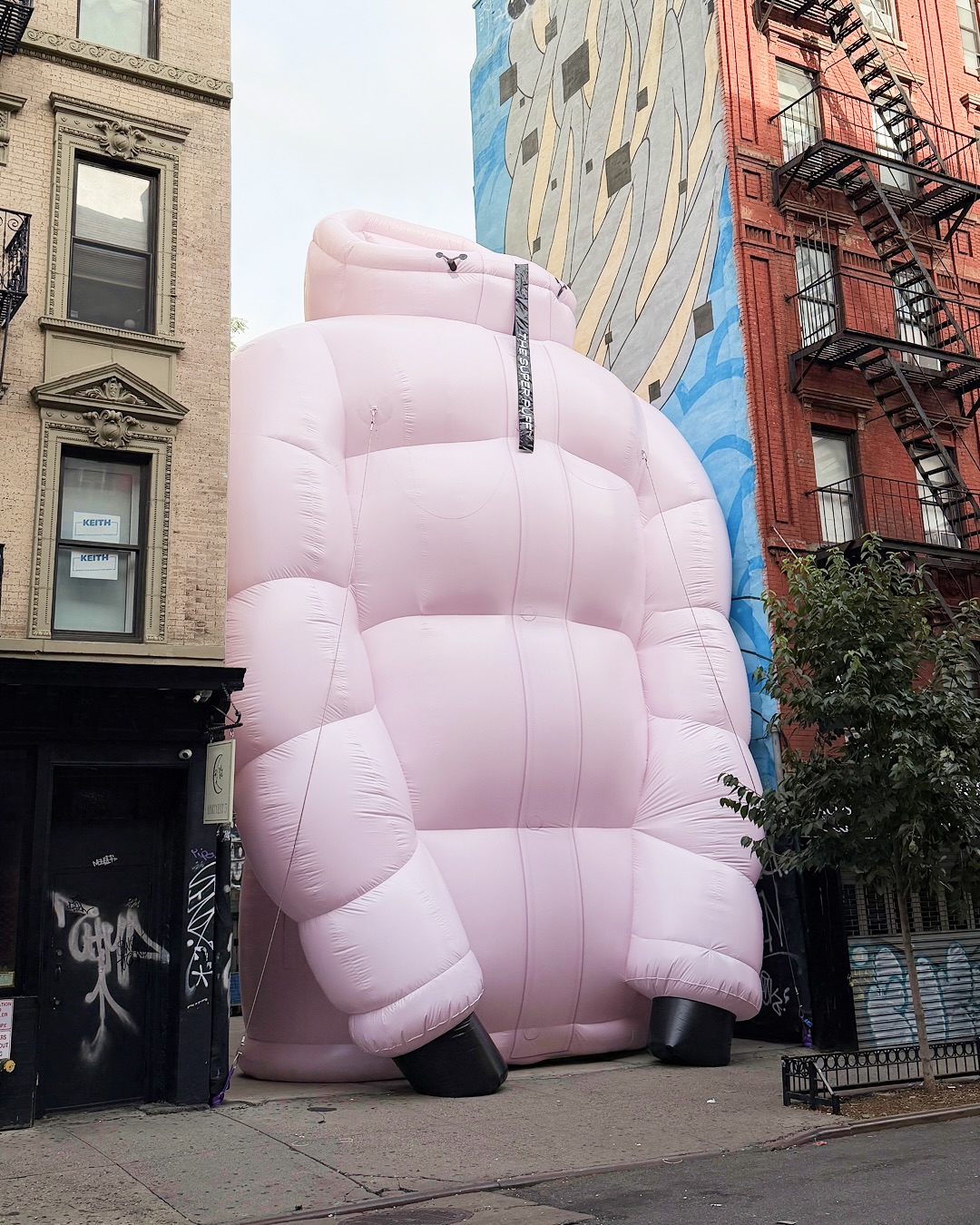 An enormous pale pink Super Puff jacket fills a narrow gap between two New York buildings, dwarfing people beside it.