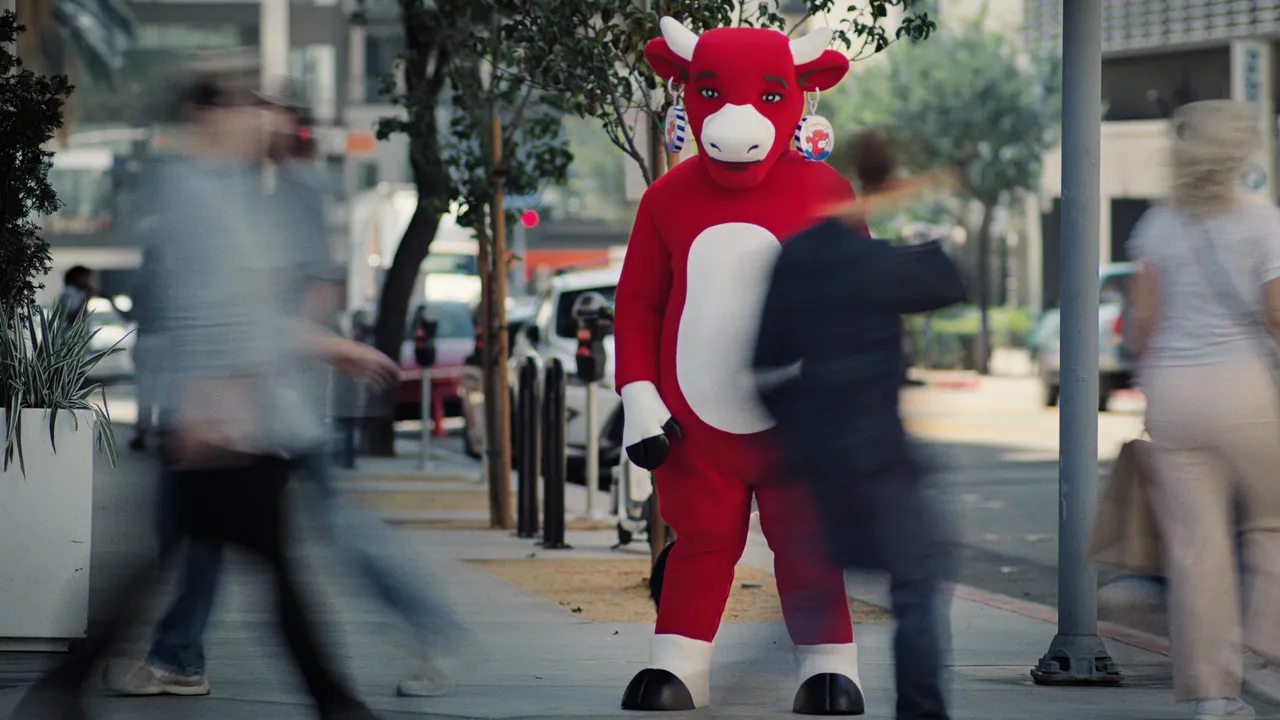 An unsmiling red Laughing Cow character stands in a city street.
