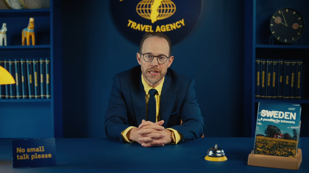 A suited host sits at a blue travel-agency desk beside a Sweden brochure and a sign reading “No small talk please”.