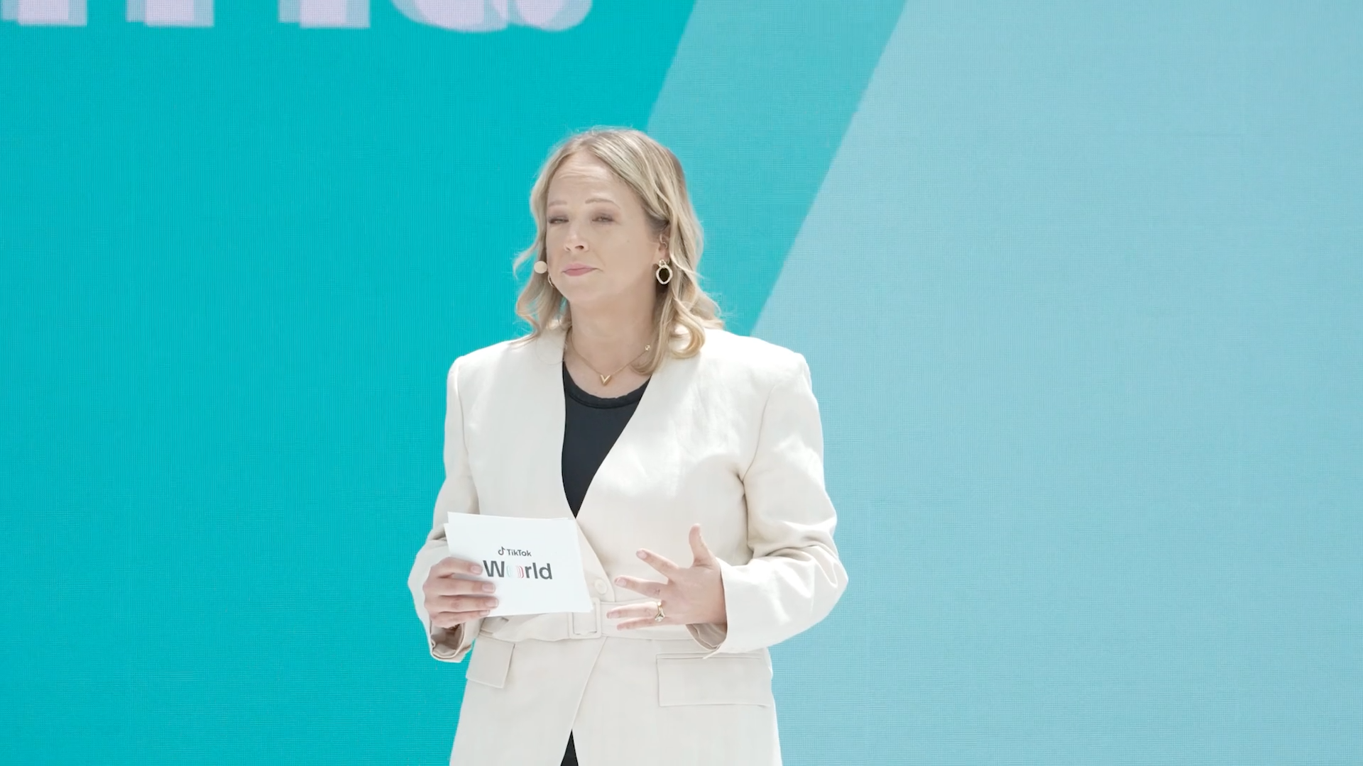 A TikTok World presenter in a white suit holds a branded cue card on a turquoise stage.