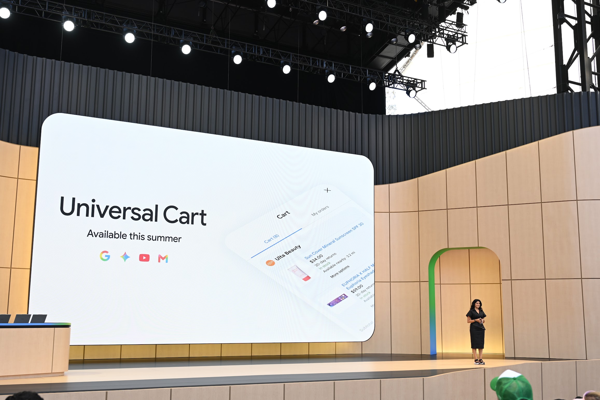 A presenter stands on the Google I/O stage beside a Universal Cart launch slide, which shows a summer rollout and an example shopping cart.