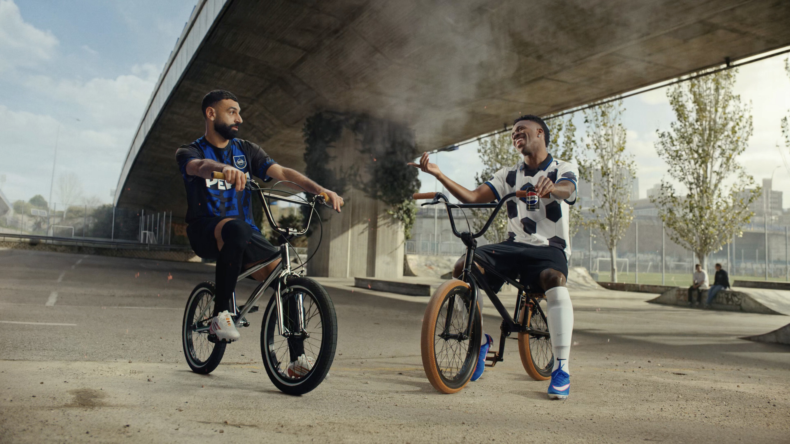Mohamed Salah and Vini Jr. sit on BMX bicycles beneath a concrete flyover in Pepsi’s Football Nation campaign.