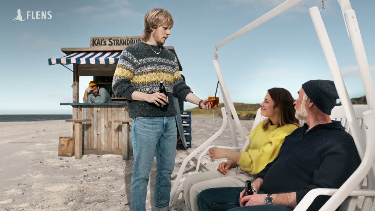 A server holding an orange drink approaches two people in a beach chair outside a seaside kiosk, with the FLENS mark above.