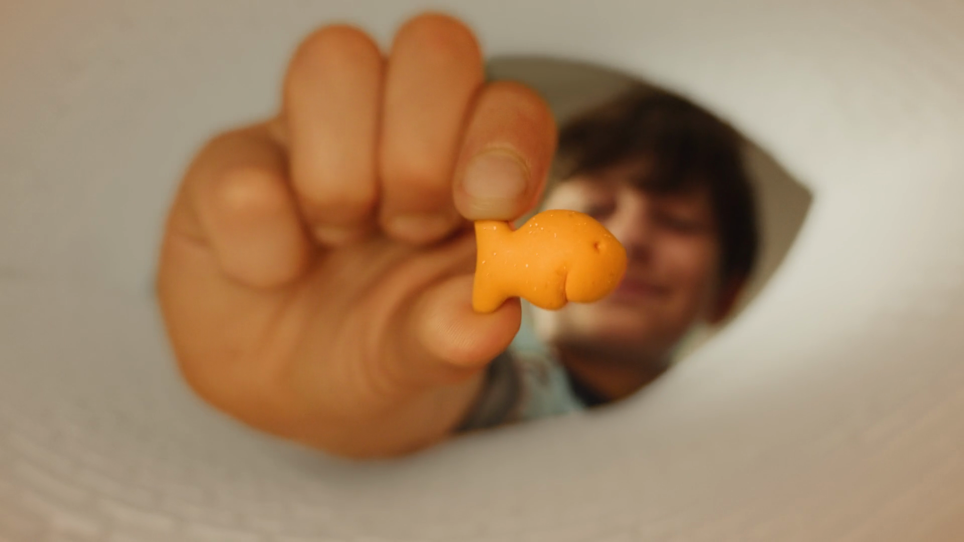 A hand holds a smiling Goldfish cracker close to the camera, with a young person’s face softly out of focus behind it.