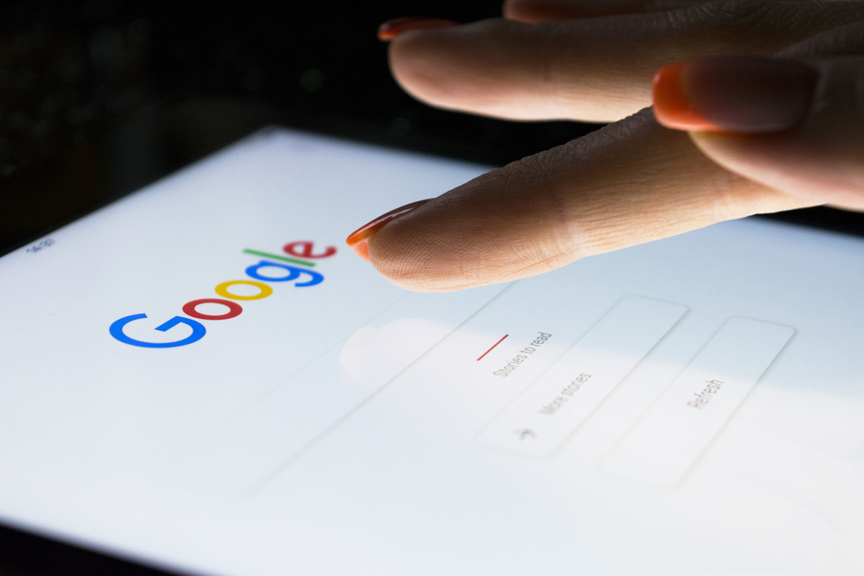 A finger uses Google Search on a white tablet held diagonally.