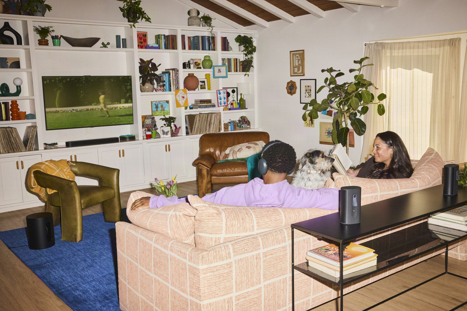 Two people and a dog sit on a sofa in a room with a Sonos soundbar and rear speakers; one listener wears headphones.